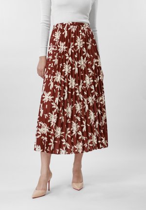 Femme portant une jupe midi plissée couleur rouille avec des motifs floraux blancs, un haut blanc à manches longues et des chaussures à talons hauts couleur nude.