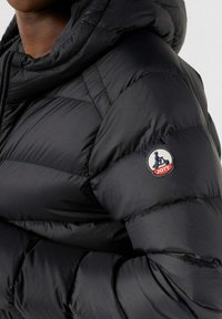 Zwarte pufferjack met capuchon, met een doorgestikte textuur en een rond patchlogo op de mouw. Het materiaal lijkt glad en glanzend.