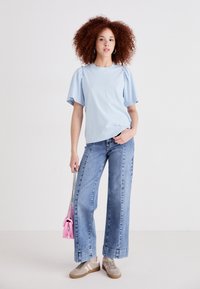 T-shirt bleu clair à manches courtes avec des détails froncés, associé à un jean effilé taille haute en denim clair. Baskets marron et petit sac à main rose.