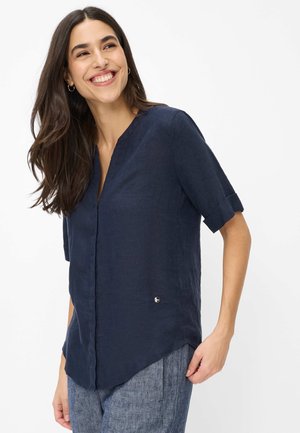 Femme souriante, portant un chemisier bleu marine à manches courtes avec un col en V et des poignets retroussés, assorti à un pantalon bleu texturé.