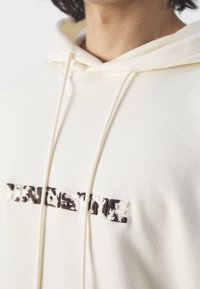 Personne portant un sweat à capuche crème avec un nom de marque à texture imprimé vache sur la poitrine et des cordons suspendus à la capuche.