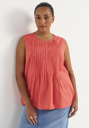 Kobieta z włosami zaczesanymi do tyłu, nosząca złote kolczyki-koła, bezrękawnik z plisowanego koralowego materiału, niebieską spódnicę jeansową i złote pierścionki na palcach.