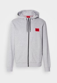 Sweat à capuche gris clair zippé avec cordons noirs et un patch rouge marqué "HUGO" sur la poitrine et à l'intérieur du col.
