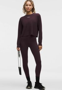 Giovane in maglia lunga viola scuro e leggings sta in piedi, tenendo una corda per saltare nera, indossando scarpe e calze sportive nere.