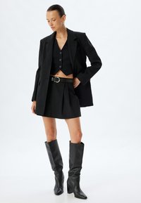 Blazer nero su misura sopra un gilet nero corto, abbinato a una gonna alta in vita e stivali di pelle fino al ginocchio. Silhouette elegante e aderente.