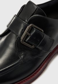 Fekete, texturált felületű bőr cipő, amely hangsúlyos pánttal és fém csattal díszített, valamint kontrasztos bordó talppal rendelkezik.