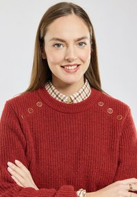 Pull rouge tricoté avec une texture côtelée, orné de trois boutons décoratifs sur l'épaule. En dessous, un col de chemise à carreaux.