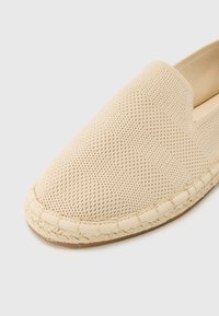 Beige espadrille met een gestructureerde netstof bovenkant, vlak ontwerp en gevlochten jute zool. Ronde teen en elastische zijpaneel voor gemakkelijk aan- en uittrekken.