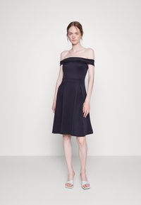 Vestido azul-marinho com ombros à mostra, corpo ajustado e saia rodada, apresentando pregas e uma textura de tecido suave. Usado com sandálias de cor clara.