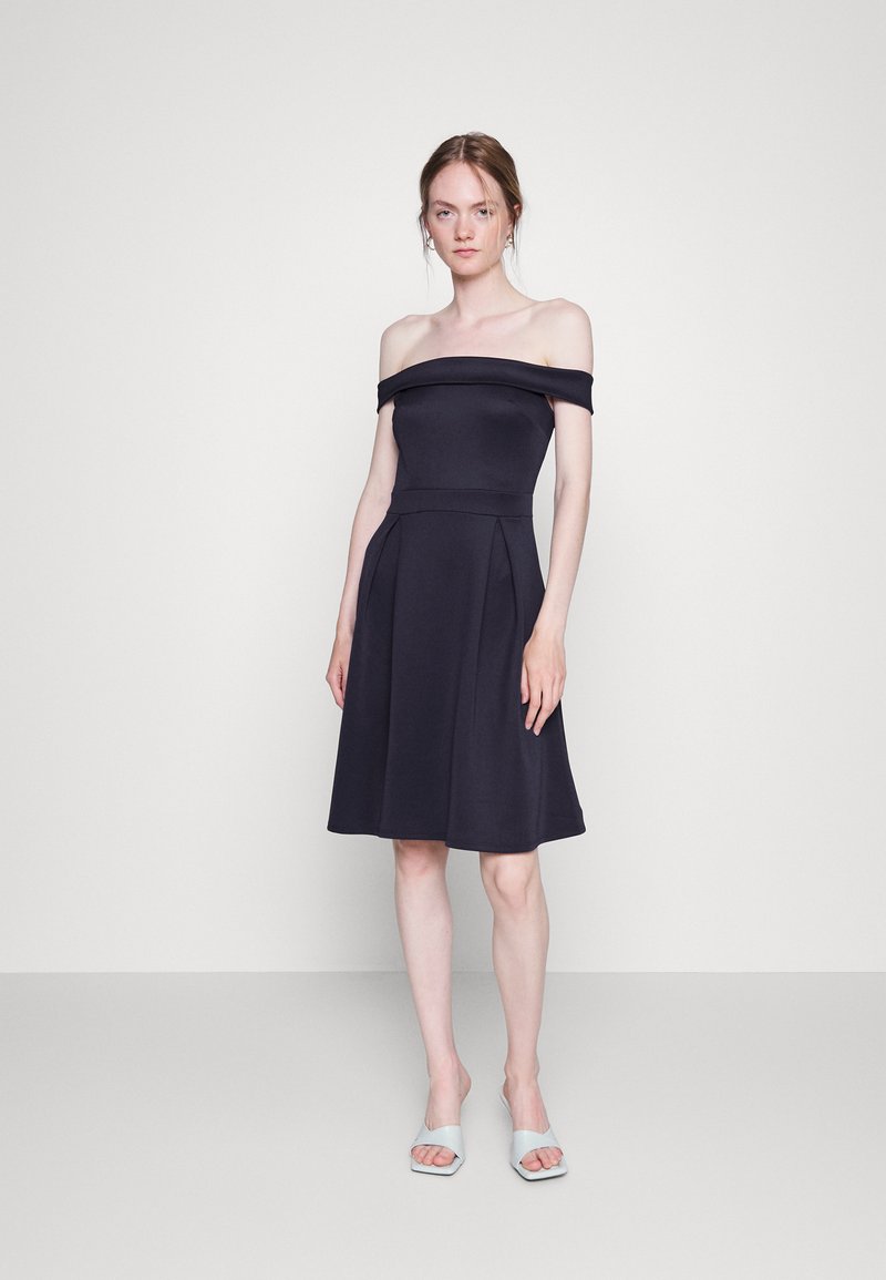 Vestido azul-marinho com ombros à mostra, corpo ajustado e saia rodada, apresentando pregas e uma textura de tecido suave. Usado com sandálias de cor clara.