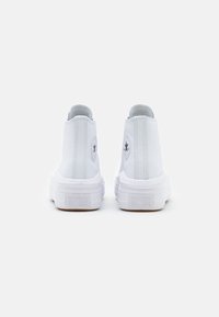 Białe sneakersy za kostkę z gładką skórzaną powierzchnią, zaokrąglonym noskiem i wyraźną gumową podeszwą. Na pięcie znajduje się okrągła naszywka z logo.