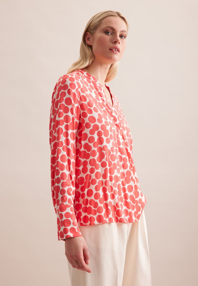 Blusa a maniche lunghe corallo con grandi pois rossi, scollatura a V, tessuto morbido e vestibilità relaxed. Abbinata a pantaloni chiari.