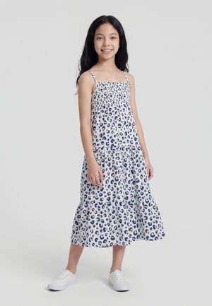 Young girl with long dark hair wearing a sleeveless white dress with blue leopard print and white sneakers, standing and smiling.