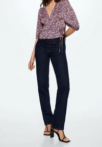 Donna che indossa una blusa avvolgente floreale viola, jeans a gamba dritta blu scuro e sandali neri con tacco alto, in piedi contro uno sfondo semplice.
