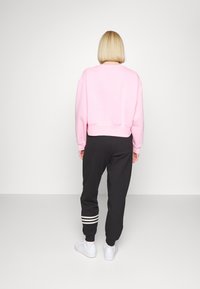 Žena s krátkými blond vlasy, oblečená v růžové mikině, černých teplákových kalhotách s bílými pruhy u kotníků a bílých teniskách, stojící u bílé zdi.