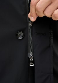 Hand, die den Reißverschluss einer schwarzen Jacke hält, mit sichtbarem Knopf und angenähtem Knopfloch in der Nähe.