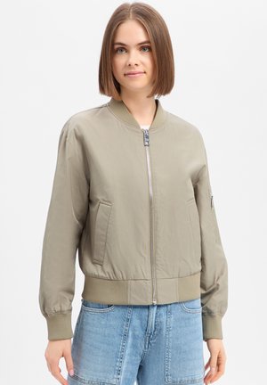 Frau mit glatten braunen Haaren, trägt eine hellbeige Bomberjacke mit Reißverschluss und hellblaue Jeans vor einem schlichten Hintergrund.