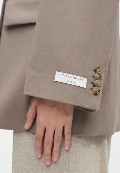 Μπεζ ύφασμα σακακιού με απαλό υφή, που διαθέτει τέσσερα καφέ κουμπιά στον καρπό και μια λευκή ετικέτα που γράφει "TIGER OF SWEDEN 1903."