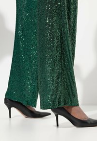 Groene pailletten broek met wijde pijpen, gecombineerd met zwarte stiletto heels. De stof glinstert met een gestructureerd, geribbeld ontwerp en een gladde afwerking van de schoenen.