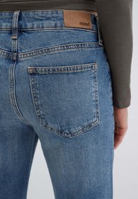 Jeans in denim blu con una tasca posteriore visibile, etichetta in pelle marrone in vita, cuciture a contrasto e una leggera texture sbiadita.