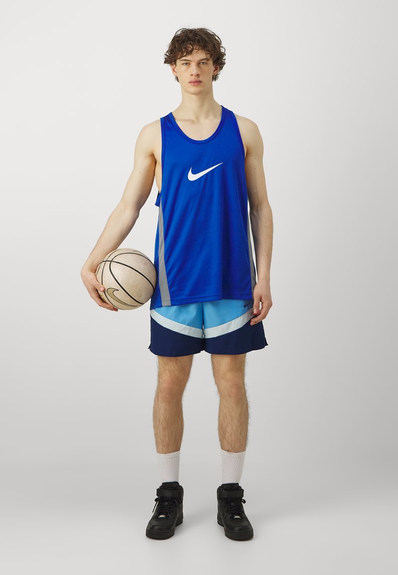 Canotta da basket in mesh blu con logo Nike bianco, abbinata a pantaloni corti azzurri e blu navy. Il modello tiene in mano un pallone da basket, indossando sneakers nere.