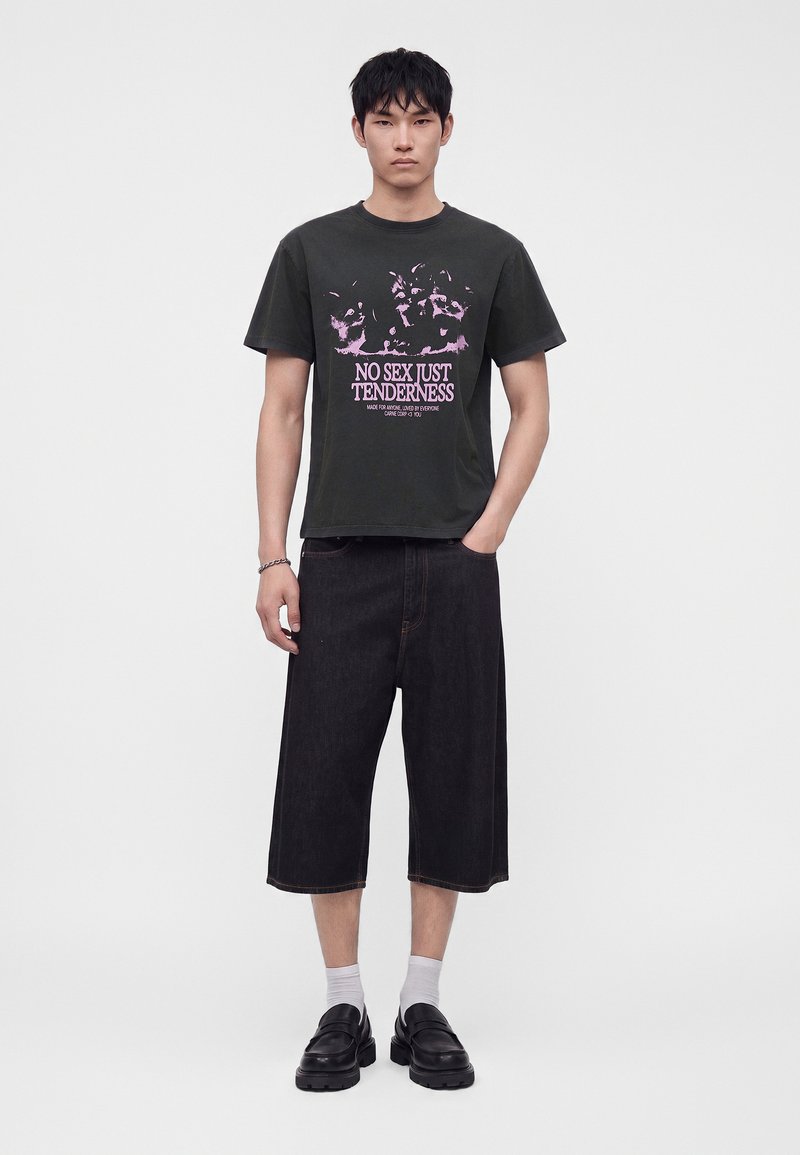 Νέος άνδρας που φοράει μαύρο T-shirt με ροζ κείμενο "No sex just tenderness,", μαύρα φαρδιά σορτς, λευκές κάλτσες και μαύρα παπούτσια, στέκεται μπροστά σε απλό φόντο.