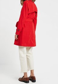 Trench rojo con cintura ceñida con cinturón, hombreras y una abertura trasera, combinado con pantalones blancos cortos y zapatos marrones.