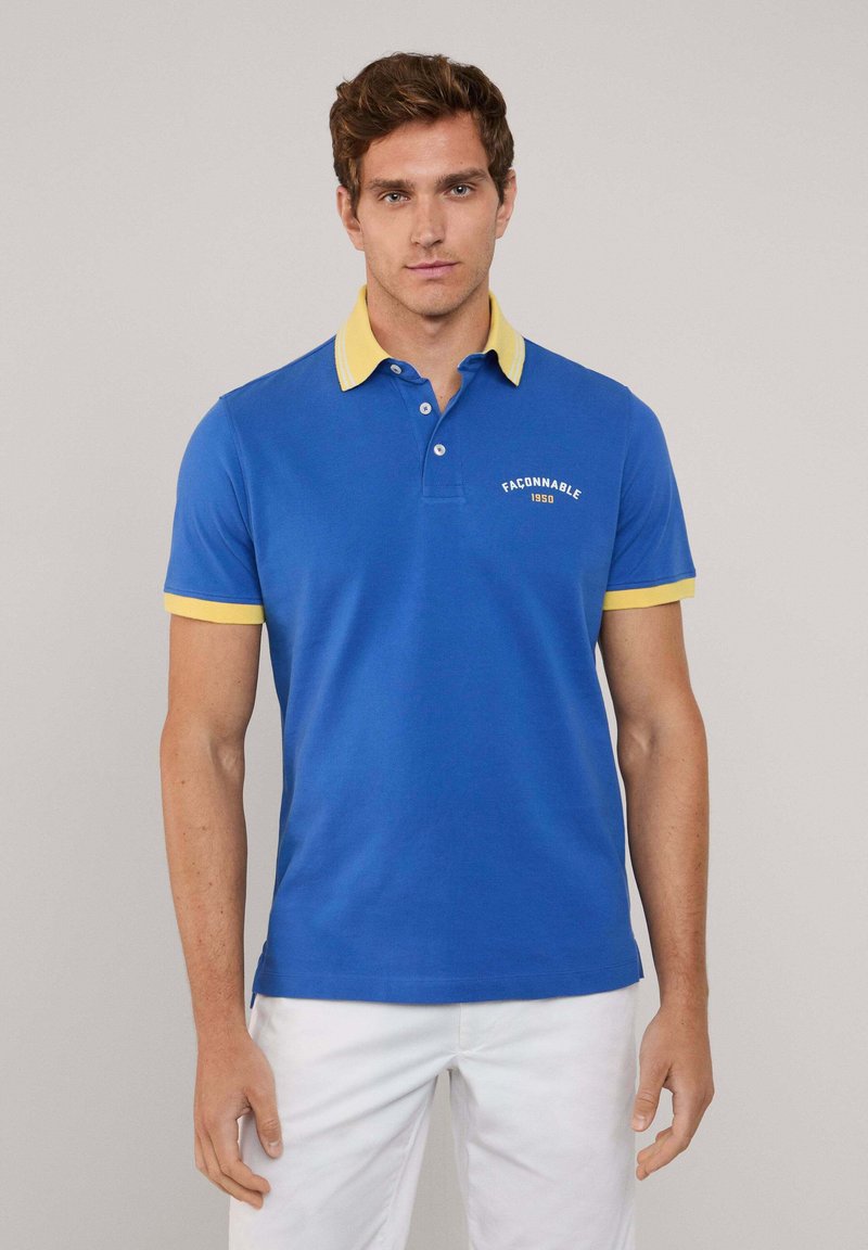 Niebieska koszulka polo z żółtym kołnierzykiem i akcentami na rękawach, z trzema guzikami przy dekolcie i haftowanym logo po lewej stronie na klatce piersiowej.