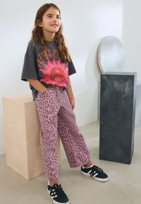 T-shirt grafico grigio con design di sole rosa, abbinato a pantaloni a stampa leopardata rosa. Sneakers nere completano l'outfit. Elementi di sfondo minimalisti.