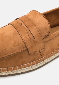 Brown suede loafers with a flat weave jute sole, featuring a decorative strap across the top and visible stitching details.