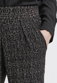 Czarne i srebrne spodnie w wzory z teksturowanej tkaniny, z płaskim przodem i bocznymi kieszeniami, zaprezentowane z częściowo widoczną czarną bluzką.