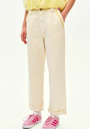 Osoba w kremowych spodniach o prostych nogawkach z żółtym falistym wzorem, żółtym swetrze i różowych trampkach, stojąca z jedną ręką w kieszeni.