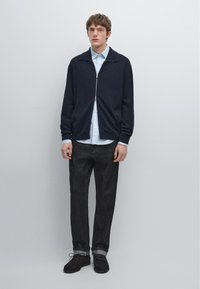 Námořnický svetr na zip přes světle modrou košili s knoflíky, doplněný o tmavé džíny a černé boty. Jednoduchý design bez výrazných vzorů.