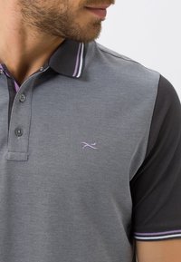 Polo gris avec des manches noires, présentant une texture unie, un col boutonné et un petit logo violet sur la poitrine. Accents de col à rayures doubles.