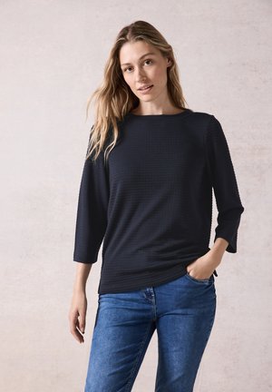 Svart texturerad långärmad topp med rund halsringning, bärs med blå jeans, har en avslappnad passform och en sidospalt.