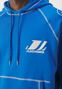 Gros plan d'une personne portant un sweat à capuche bleu avec des coutures blanches et un logo blanc J.Lindeberg sur la poitrine.