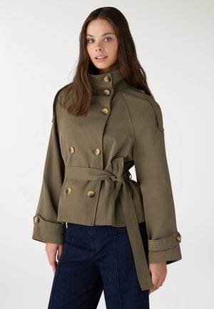 Donna con capelli castano lunghi che indossa una giacca doppiopetto verde oliva con grandi bottoni e cintura annodata in vita, abbinata a pantaloni scuri.