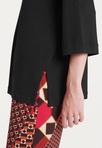 T-shirt nera con maniche larghe, caratterizzata da spacchi laterali. Abbinata a pantaloni a motivi rossi e marroni che mostrano disegni geometrici.