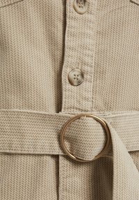Tessuto beige testurizzato con motivo a spina di pesce. Presenta bottoni e una cintura con una fibbia circolare dorata. Design di lunghezza media.