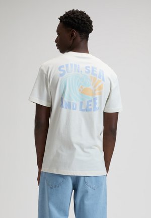 Mees seljaga, kellel on seljas valge T-särk ja sinised teksad, T-särgi tagaküljel on lainetus, päike ja tekst "PÄIKE, MERE JA LEE" pastellsetes toonides.