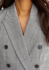 Blazer gris de doble botonadura con un patrón a cuadros, con botones negros y un acabado texturizado. Las solapas anchas realzan el diseño estructurado.