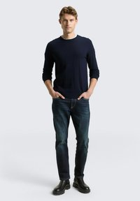 Marineblauwe geribde trui met lange mouwen, gecombineerd met donkerblauwe jeans en zwarte enkellaarsjes. Model staat met handen in zakken, neutrale achtergrond.