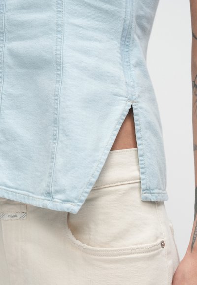 Κοντινό πλάνο ανοιχτό μπλε τζιν κορυφής με πλευρικό σχίσιμο πάνω από κρεμ παντελόνι ψηλής μέσης, με εμφανές τατουάζ στο δεξί χέρι.