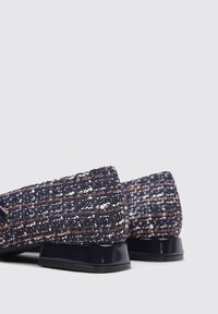 Chaussures plates en tissu bleu tissé avec une finition texturée, dotées d'un petit talon noir en vernis. De subtiles taches orange et blanches rehaussent le design.