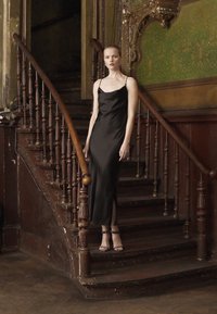 Black silk slip dress with thin straps, featuring a side slit, worn by a model on a wooden stairway with ornate railings and dark walls.
