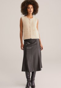 Gilet lavorato a maglia beige e morbido con grandi bottoni e tasche, abbinato a una gonna midi in pelle nera e stivali in pelle al ginocchio.