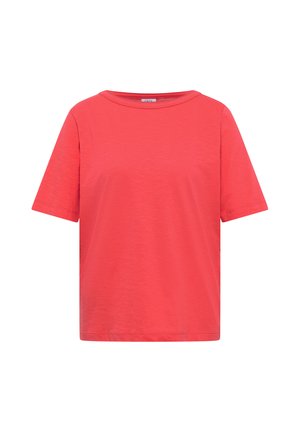 Femme souriante, de profil, portant un t-shirt corail vif à manches courtes et un jean bleu sur un fond beige uni.