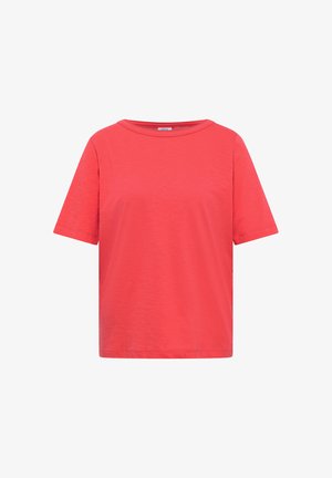Femme souriante, de profil, portant un t-shirt corail vif à manches courtes et un jean bleu sur un fond beige uni.