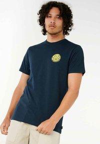 Marineblå T-shirt lavet af bomuld, med et cirkulært gult grafisk logo på brystet. Kortærmet og med rund hals.