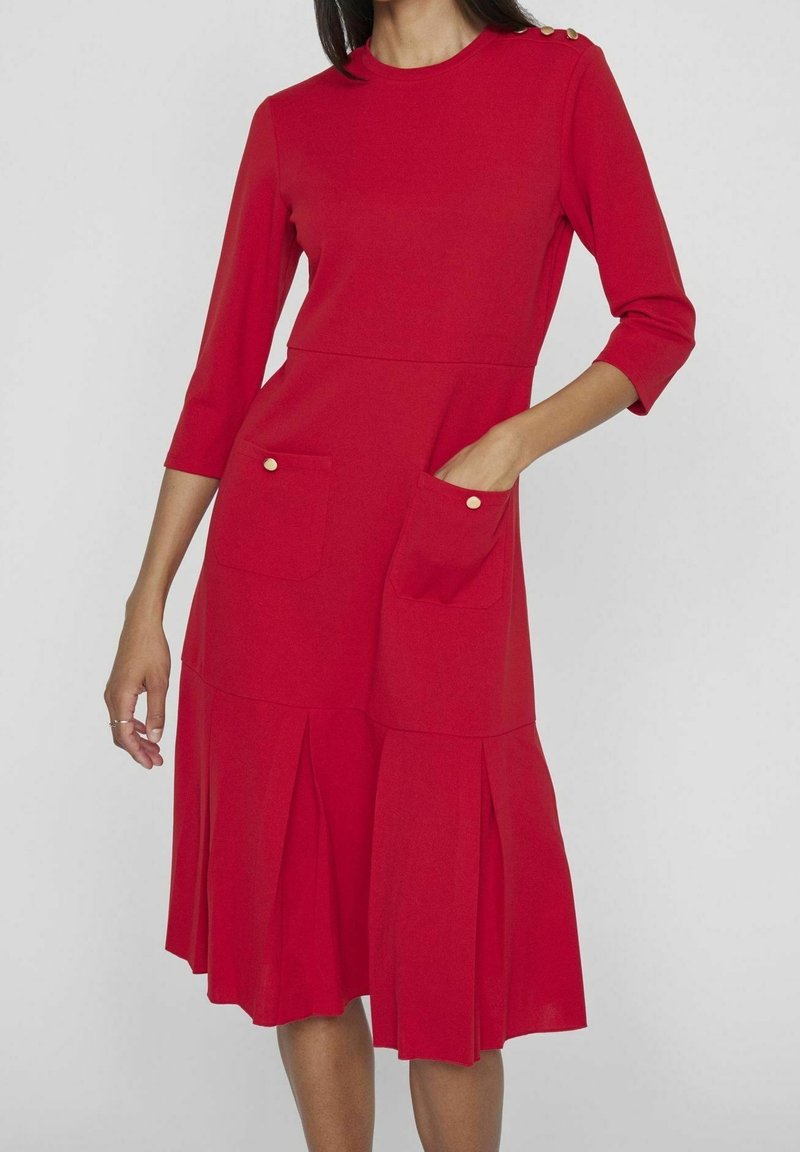 Femme portant une robe rouge longueur genou avec des manches trois-quarts, des boutons dorés sur l'épaule et deux poches avant, une main dans la poche.